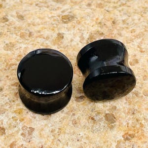 Peut inclure: Deux bouchons d'oreille noirs sont présentés. Les bouchons ont une forme arrondie et évasée avec une finition lisse et brillante. Les bouchons sont faits d'un matériau sombre et solide, et sont posés sur une surface texturée brun clair.