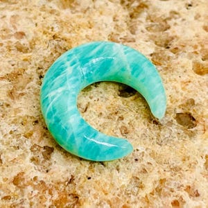 Puede incluir: Una piedra preciosa pulida en forma de media luna, en tonos turquesa y blanco. La piedra tiene un acabado liso y brillante y un patrón natural veteado. La forma curva recuerda a una luna. La gema se muestra sobre una superficie texturizada y beige.