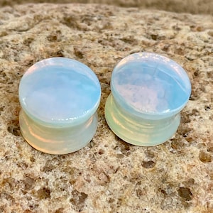 Könnte beinhalten: Zwei opaleszierende Ohrstöpsel mit einem hellblauen und weißen Farbverlauf. Die Stöpsel haben eine abgerundete Oberseite und eine ausgestellte Basis. Sie liegen auf einer strukturierten, hellbraunen Oberfläche.