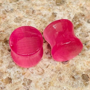 May include: Two bright pink ear gauges. One is a flat disc shape, and the other is a flared plug shape. The gauges are made of a smooth, glossy material. The background is a light brown, textured surface.