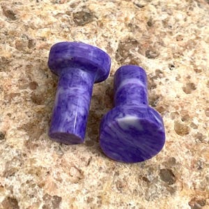 May include: Two purple and white marbled stone bottle stoppers. The stoppers have a cylindrical stem and a rounded top. The color is a deep purple with white marbling throughout the stone.