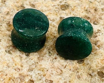 May include: Two dark green stone ear plugs with a flared design. The plugs have a smooth, polished surface and a slightly rounded shape. The plugs are displayed on a textured, light brown surface.