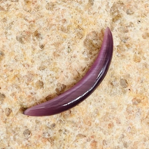 May include: A curved, polished purple gemstone, possibly a pendant or bead, with a smooth, glossy finish. The gemstone has a crescent shape and a gradient of purple hues, from light to dark. The object is set against a textured, light brown surface.