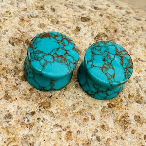 May include: Two turquoise stone ear plugs with brown veining. The plugs are cylindrical with a flared base. The stone has a polished, smooth surface. The plugs are displayed on a textured, light brown surface.