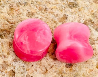 Pode incluir: Dois tampões de ouvido rosa com efeito marmorizado. Um é um cilindro arredondado, e o outro tem uma forma mais orgânica e alargada. Os tampões estão sobre um fundo texturizado e bege.
