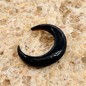 May include: A glossy black crescent-shaped object, possibly jewelry or a decorative element. The object has a smooth, curved surface and a dark, reflective appearance. The background is a textured, light brown surface.