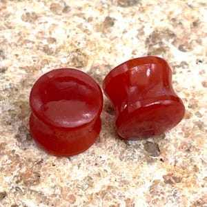 May include: Two polished, red-orange ear plugs. The plugs have a flared design, with a wider base and top, and a narrower center. The material appears smooth and translucent, with a glossy finish. The plugs are displayed on a textured, light brown surface.
