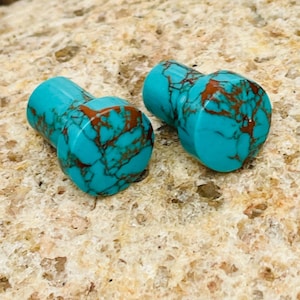 May include: Two turquoise plugs with brown veining. The plugs are cylindrical with a rounded face. The turquoise color is bright and the brown veining creates a marbled effect. The plugs are displayed on a light brown, textured surface.