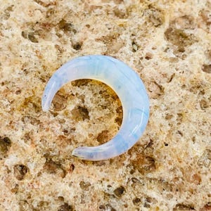 Puede incluir: Un colgante de luna opalescente en forma de media luna. El colgante es de color azul pálido y blanco, con un aspecto translúcido. La luna está colocada sobre una superficie texturizada de color marrón claro. El colgante es probablemente una joya.