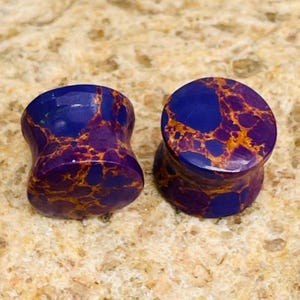 May include: Two purple and blue stone ear plugs with a marbled pattern. The plugs have a flared design and a polished finish. The stone has a mix of purple, blue, and gold colors.