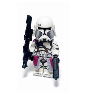 Puede incluir: Una figura de acción blanca y gris de un soldado con casco, que sostiene un rifle negro y una pistola negra. La figura tiene un detalle rosa y gris en forma de falda y correas marrones.