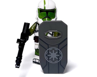 May include: A Lego minifigure of a soldier in white and green armor, holding a black rifle and a gray shield with a star emblem. The figure is standing against a white background.
