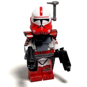 May include: A red, white, and black action figure of a soldier with a helmet, armour, and two black blasters. The figure has a grey accessory attached to the helmet. The figure is a collectible toy.