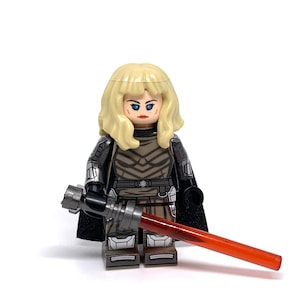 May include: A Lego minifigure of a female character with blonde hair, blue eyes, and a stern expression. She wears a detailed gray and black outfit with a black cape and holds a silver and black lightsaber with a red blade. The figure is on a white background.