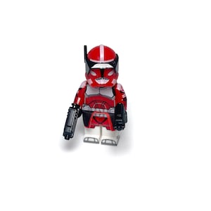 Può includere: Figura giocattolo rossa e bianca di un soldato con elmetto e accessori neri. La figura tiene un'arma nera nella mano sinistra e ha un accessorio nero sul lato destro. La figura è su una superficie bianca.