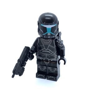 May include: A black plastic minifigure wearing a helmet with a light blue visor and silver detailing. The torso and legs feature printed silver armor patterns. The figure holds a black plastic blaster accessory in its right hand.