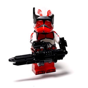 May include: A red, white, and black toy figurine of a soldier holding a black weapon. The figurine has a helmet with red and white markings and is wearing red and white armour. The toy is standing on a white surface.