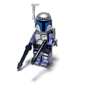 Puede incluir: Una minifigura Lego Star Wars Jango Fett. La figura tiene un casco plateado con detalles azules, un torso azul y gris, y piernas azules y grises. Sostiene dos blasters negros y tiene una mochila propulsora plateada.