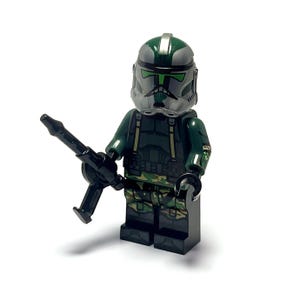 May include: A Lego minifigure of a soldier in a green and gray helmet and uniform, holding a black weapon. The figure has camouflage pants and is standing on a black base. The background is white.
