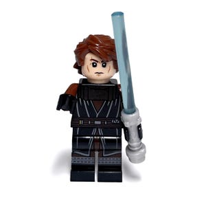 May include: A Lego minifigure of a character with brown hair, wearing a black and brown outfit, and holding a blue lightsaber. The figure has a stern expression and is posed against a white background.