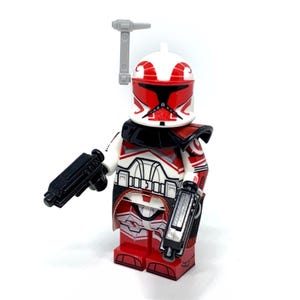 May include: A red, white, and black Lego minifigure of a clone trooper with two black blasters and a grey rangefinder. The minifigure has a white helmet with red markings and armour with red and white details.