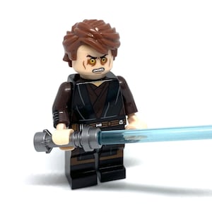 Puede incluir: Una minifigura de Lego de un personaje con cabello castaño y una expresión severa, vistiendo un atuendo marrón oscuro y negro. La figura sostiene un sable de luz azul, con una empuñadura plateada. El fondo es blanco.