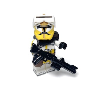 Puede incluir: Minifigura Lego de un soldado clon de Star Wars. La figura es principalmente blanca y amarilla con detalles en negro, lleva un casco y sostiene un rifle bláster negro. El fondo es blanco.