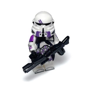 May include: A white and purple action figure of a soldier holding a black rifle. The figure has a white helmet with purple accents, and purple details on the shoulders and torso. The figure is standing on a white surface.