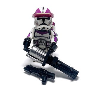 Könnte beinhalten: Eine weiße und lila Lego-Minifigur eines Klonkriegers mit Helm und Visier. Die Minifigur hält eine silberne und schwarze Waffe und hat eine schwarze Pistole auf dem Boden. Die Figur hat ein lila und weißes Farbschema.