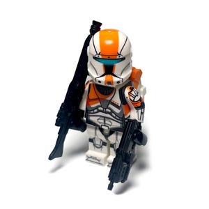 May include: A white and orange action figure with black weapons. The figure has a helmet with orange stripes and a blue visor. It is wearing armor with orange and gray accents and is holding two black guns.