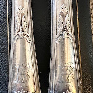 May include: Close-up of two antique silver utensils. Each utensil features an ornate, engraved design with a floral motif and the letter "B". The handles have a decorative, symmetrical pattern. The silver has a polished finish.