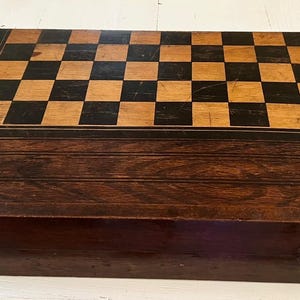 Puede incluir: Un juego de ajedrez de madera vintage con una parte superior de tablero de ajedrez con cuadrados negros y beige alternados. La caja de madera marrón oscuro tiene forma rectangular con un panel decorativo en la parte delantera y asas de metal. El tablero de ajedrez mide unos 38 cm por 38 cm.