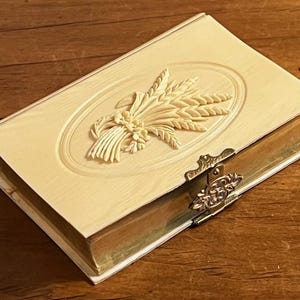 May include: Ivory-colored rectangular box with a wheat sheaf design on the lid. The box has a gold-colored metal clasp and trim. The box is likely used for storing small items and is placed on a wooden surface.