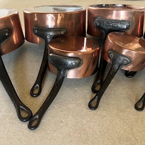 May include: A set of seven graduated copper saucepans with polished metallic surfaces and dark, textured iron handles. Each pan features a cylindrical body and a riveted handle attachment, arranged in a descending size order from left to right.