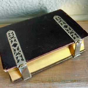 May include: A vintage book with a dark brown leather cover and gold-edged pages. Two ornate silver metal clasps secure the book, featuring a decorative cut-out design. The book rests on a wooden surface.
