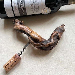 May include: A handcrafted wooden corkscrew with a dark metal screw and a cork with the word "CITADELLE" printed on it. A bottle of red wine is in the background. The corkscrew is made from natural wood.