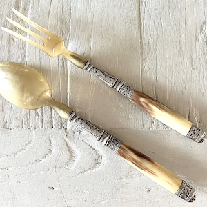 Puede incluir: Un juego de tenedor y cuchara para servir con cabezas y mangos de cuerno de color crema translúcido. Cada utensilio incluye un collar y una tapa de metal plateado, ambos grabados con intrincados patrones florales y geométricos.