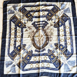 May include: A square silk scarf featuring a dark blue border and a central cream-coloured field. The design displays a brown horseshoe surrounded by white daisies and foliage, framed by blue leather-like straps, gold rings, and tassels.