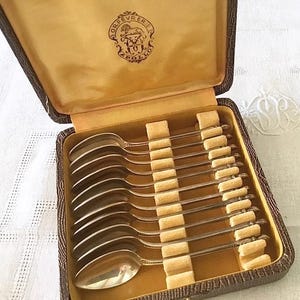 May include: A set of silver spoons in a brown case with a yellow interior. The spoons are arranged neatly in the case, with each spoon separated by a beige divider. The case has a gold emblem on the inside of the lid.