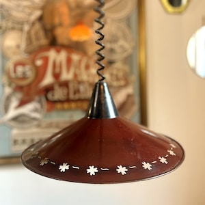 Puede incluir: Lámpara colgante de estilo vintage con pantalla granate y parte superior negra. La pantalla está decorada con recortes blancos en forma de estrella. La lámpara cuelga de un cable en espiral. El fondo presenta un cartel vintage.