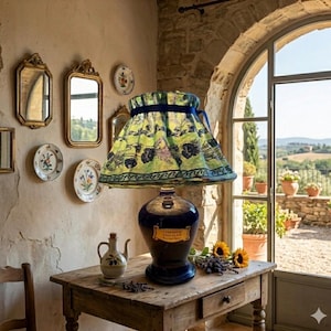 Puede incluir: Una lámpara de mesa decorativa con base azul marino y pantalla estampada. La pantalla tiene un fondo verde con diseños florales negros y un ribete de cinta azul. La lámpara está sobre una mesa de madera rústica, con una jarrita y girasoles.