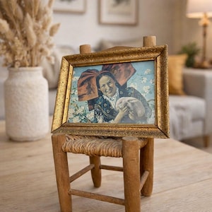 May include: A framed vintage print featuring a woman in traditional attire holding a rabbit. The print has a gold-colored ornate frame and is displayed on a small, rustic wooden chair with a woven seat. The background includes a vase with dried flowers.
