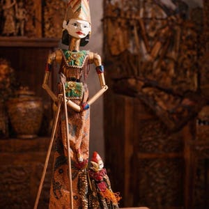 Op de afbeelding: Een houten marionet met een geschilderd gezicht, met een hoge rode hoofdtooi en kleding met patronen. De marionet houdt twee houten stokken vast en staat naast een kleinere marionet. De achtergrond bevat houten snijwerk.