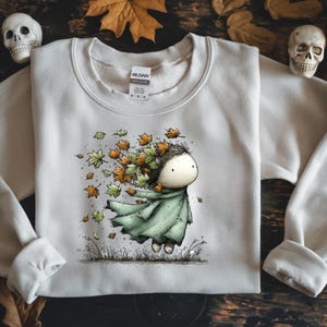May include: Cream-colored sweatshirt featuring a whimsical illustration of a figure with autumn leaves in their hair and a green cloak. The design includes falling leaves in shades of orange and green, evoking a fall theme.