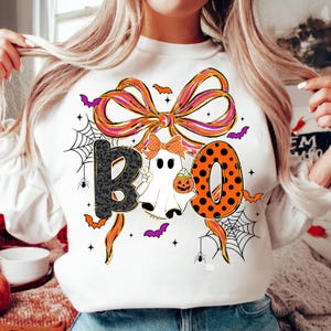 Puede incluir: Sudadera blanca de Halloween con la palabra "BOO". La "B" es negra con un estampado, la "O" es naranja con lunares negros, y la segunda "O" es un fantasma con una calabaza. Telarañas y murciélagos.