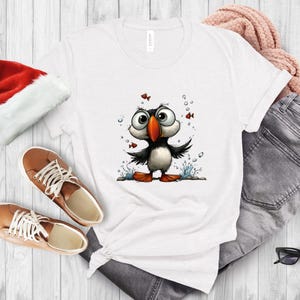 K&ouml;nnte beinhalten: Wei&szlig;es T-Shirt mit einer Cartoon-Illustration eines Papageientauchers. Der Papageientaucher hat einen schwarz-wei&szlig;en K&ouml;rper, einen orangefarbenen Schnabel und F&uuml;&szlig;e sowie gro&szlig;e Augen. Das Design umfasst Blasen und kleine rote Fische. Das Shirt wird mit braunen Schuhen und grauen Jeans gezeigt.