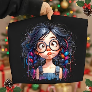 Könnte beinhalten: Schwarzes T-Shirt mit einer Cartoon-Zeichnung eines Mädchens mit runder Brille, Sommersprossen und blauem Haar, das zu Zöpfen mit Regenbogenakzenten geflochten ist. Das Mädchen hat einen leicht verwirrten Gesichtsausdruck und trägt eine Jeans-Latzhose.