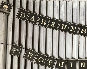 May include: Black and white banner with the text "DARKNESS AND NOTHING" in gold lettering. The banner is strung on a black cord and is hanging against a white wooden background.