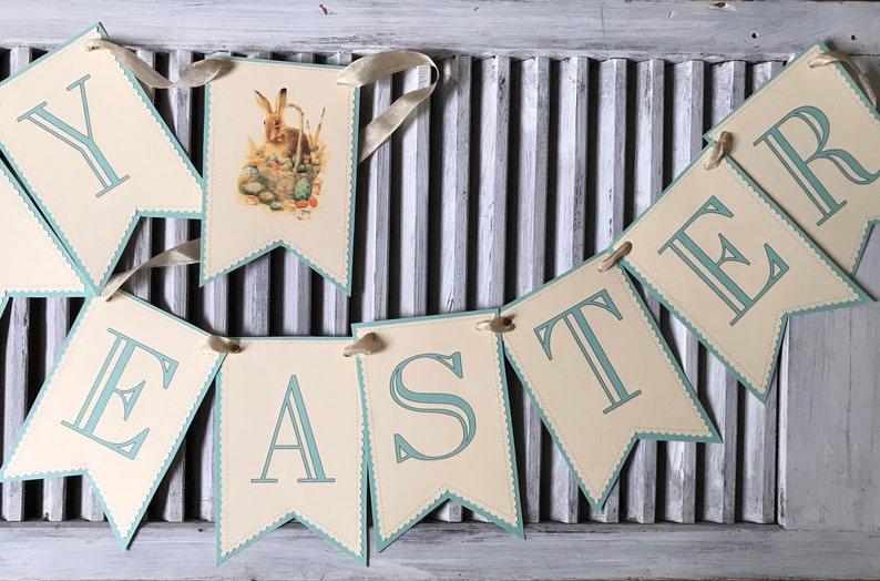 May include: A light blue and cream banner with the word "Easter" spelled out in blue letters. Each letter is on a separate flag with a scalloped edge. The flag with the letter "A" has a picture of a rabbit with a basket of eggs.