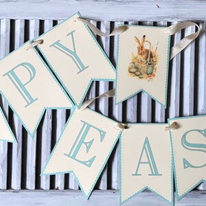 May include: A banner with the words "Happy Easter" in blue lettering on a cream background. The banner is strung with white ribbon and features a small illustration of a bunny rabbit.